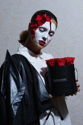 Model in a white porcelain mask with red petals, holding a black hatbox of red roses
