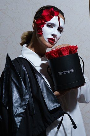 The masked model cradles the black hatbox of red roses against a marble wall