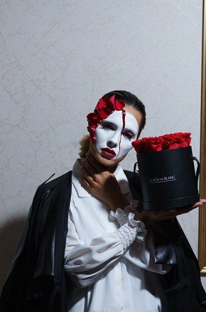 Close portrait of the masked model with her hand at her throat beside the hatbox