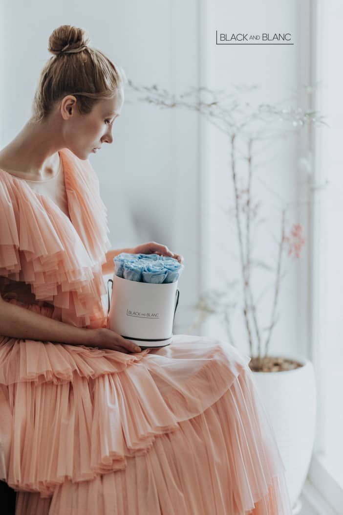Model in a peach tulle dress looks down at a white hatbox of blue roses