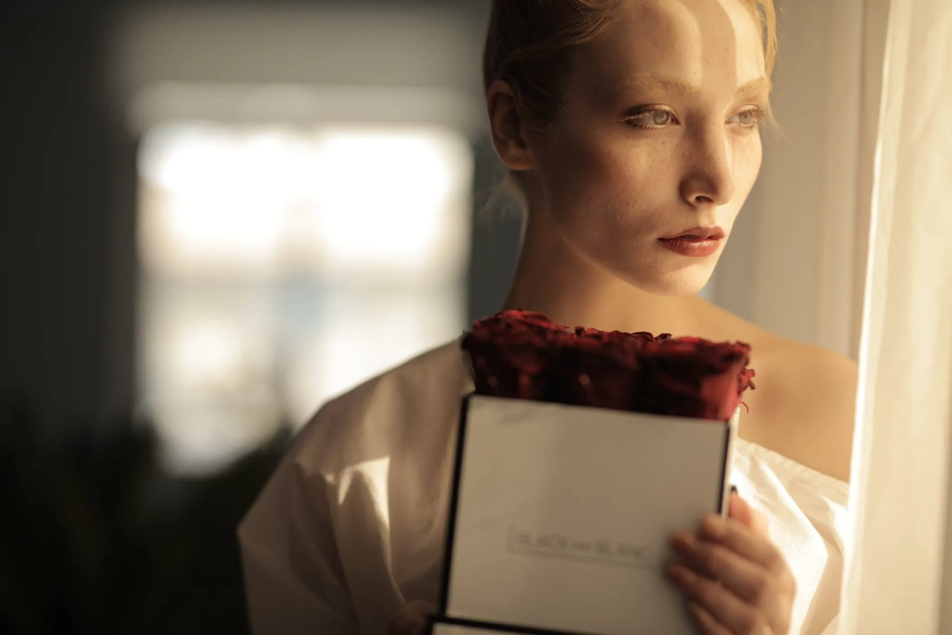 Model by a sunlit window holding a white box of deep red roses